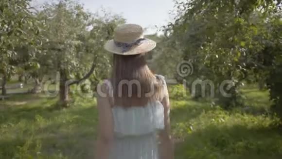 在绿色的夏日花园中漫步的迷人的年轻女子戴着草帽和白色长裙免费视频的预览图