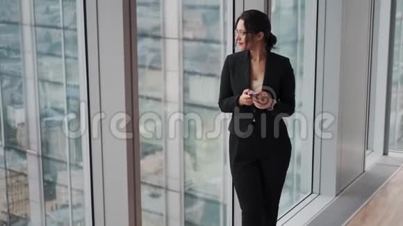 女孩站在一座摩天大楼高层的窗户旁边一位拥有智能手机的漂亮女商人的肖像视频的预览图