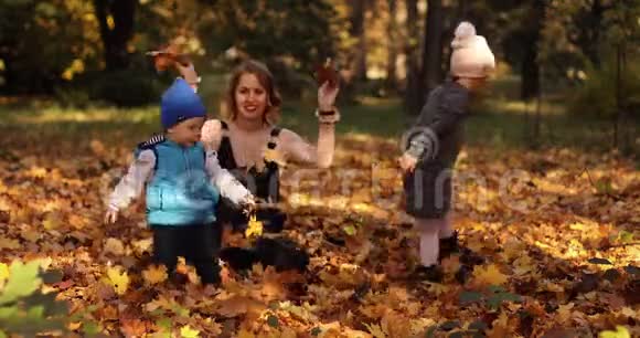 美丽的年轻母亲和两个孩子在树叶上享受阳光明媚的秋日视频的预览图