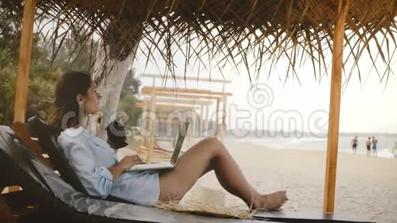 侧面观看成功的快乐自由职业者妇女与饮料享受工作移动使用笔记本电脑在躺椅海滩视频的预览图