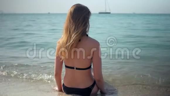 小女孩穿着黑色比基尼在阳光明媚的日子里坐在海水中的沙滩上转着身子视频的预览图