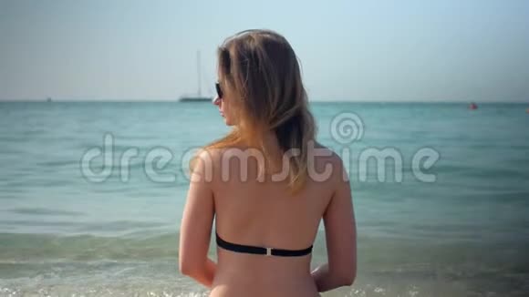 苗条的比基尼女郎正坐在沙滩上在阳光明媚的日子里看着大海视频的预览图