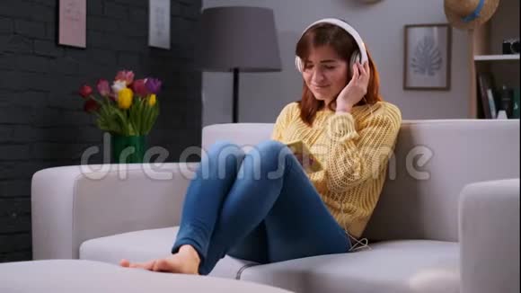 年轻迷人的女士坐在沙发上通过大白耳机和智能手机听音乐视频的预览图