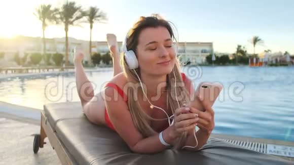穿着红色比基尼和白色耳机的可爱女孩听着智能手机上的音乐一边躺在躺椅上唱歌视频的预览图