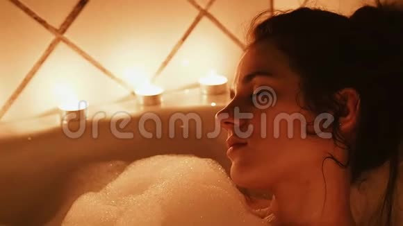 美丽的黑发女人躺在浴室里泡沫带有蜡烛的浪漫环境视频的预览图