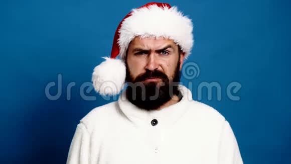 戴着新年帽子的大胡子男人在蓝色背景上扬起眉毛蓝色背景的滑稽圣诞老人概念视频的预览图