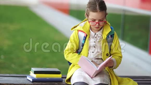 穿着黄色夹克的女学生坐在长凳上读课本在学校休息回到学校视频的预览图