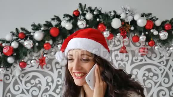 戴着圣诞帽的开朗深色卷发女士带着兴奋的表情在电话中交谈真诚地微笑模仿空间视频的预览图
