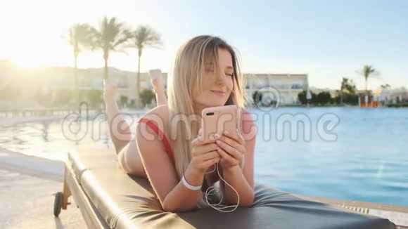 漂亮的金发女人穿着红色泳衣摇头一边听着智能手机里的音乐一边躺在躺椅上视频的预览图