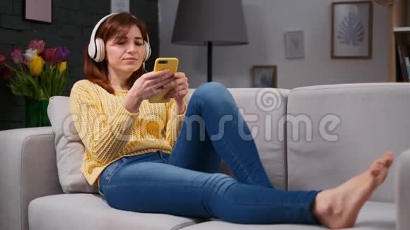 戴耳机的可爱女孩躺在客厅的沙发上听音乐和在手机上浏览的画像视频的预览图