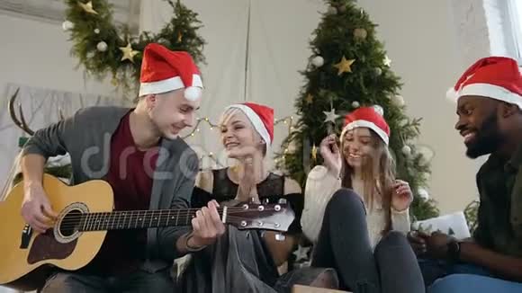 一群快乐的年轻人在家中圣诞树附近玩其中一个人在弹吉他视频的预览图