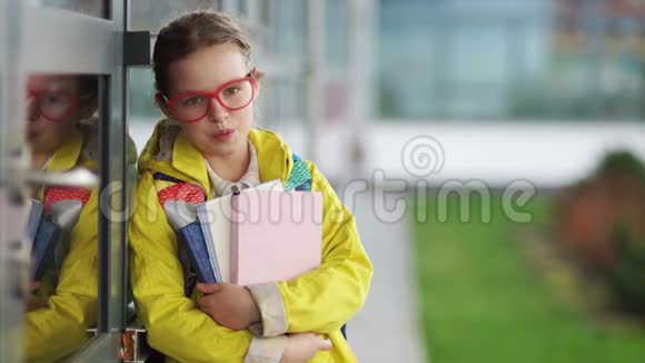 年轻女孩戴着时髦的红眼镜和一件黄色夹克手里拿着课本正在学校门口微笑视频的预览图