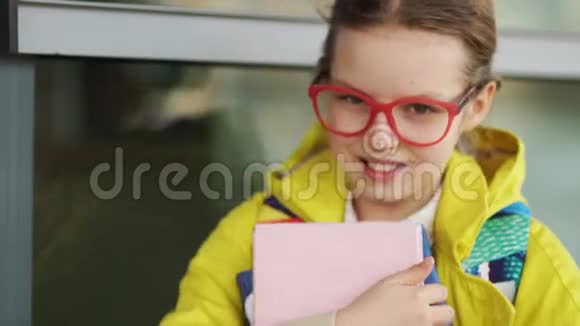 十岁的女学生高兴地对着镜头微笑戴着红眼镜的女孩和一件带课本的黄色夹克视频的预览图