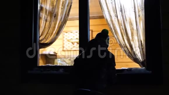 冬天的夜晚在靠近窗户的温暖明亮的房间里是一个年轻人敲着窗户在户外射击视频的预览图