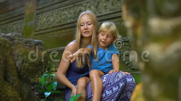 一名年轻女子和她的小儿子坐在乌布德猴子森林生态公园的楼梯上的慢镜头视频的预览图