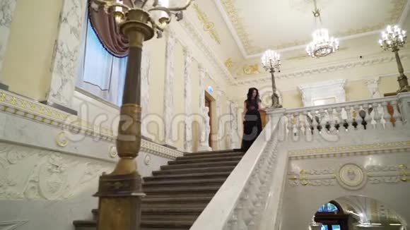 年轻的黑发女子穿着黑色晚礼服在皇宫或酒店的大理石石梯上下楼视频的预览图