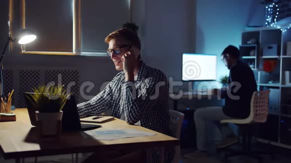 微笑的年轻人经理在黑暗的办公室工作时用手机说话视频的预览图