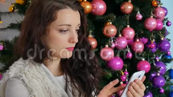 可爱的小女孩在一棵有创意的圣诞树的背景上用手机说话视频的预览图
