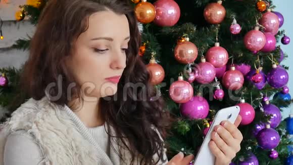 漂亮的深色头发是在一棵装饰圣诞树的背景下透过她的智能手机菜单检查视频的预览图