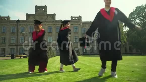 三个快乐的学生穿着黑色和红色的壁炉庆祝他们在大学大楼前毕业二视频的预览图