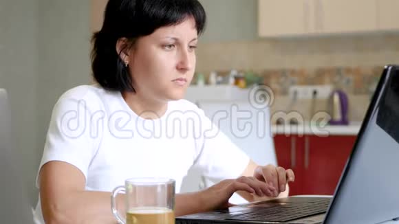年轻的黑发女人从玻璃杯里喝茶用笔记本电脑视频的预览图