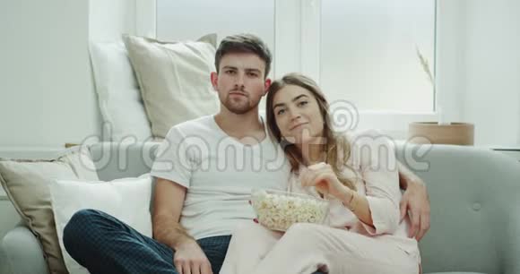 早上看电视的年轻夫妇坐在沙发上穿着睡衣吃了一大盘爆米花视频的预览图