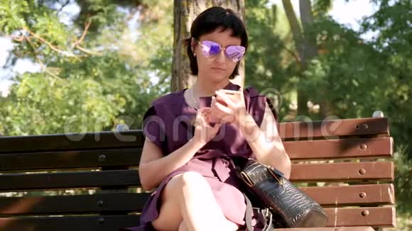 女孩坐在公园长凳上用智能手机在社交网络上交流年轻的黑发女人视频的预览图