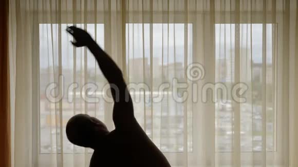 一个男人在室内做运动的剪影在背景城市街道的窗帘后面男人做热身运动视频的预览图