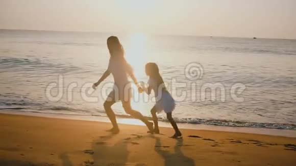 快乐美丽的年轻妈妈和小女儿牵着手一起奔跑在史诗般的夕阳海边放松视频的预览图