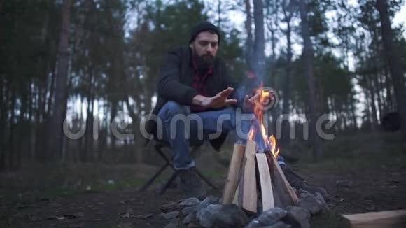 一个留着胡须的年轻人坐在椅子上在炉火旁热身森林里的火堆旁视频的预览图