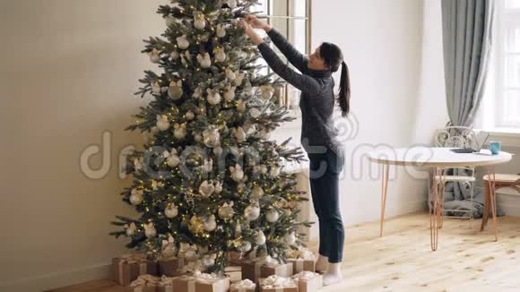 苗条的年轻女士正在装饰新年树触摸闪亮的球和灯站在室内独自在家圣诞节视频的预览图