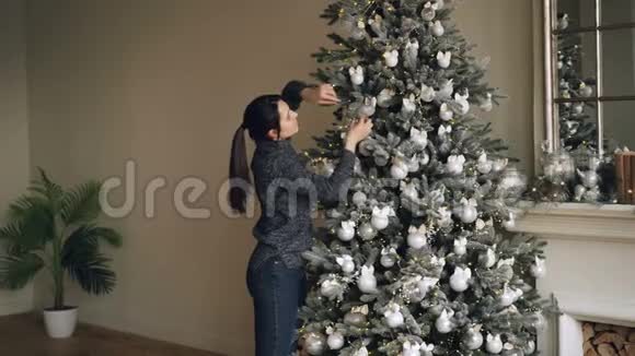 漂亮的深褐色头发的女人正在用漂亮的球和灯装饰杉树在冬天享受节日活动视频的预览图