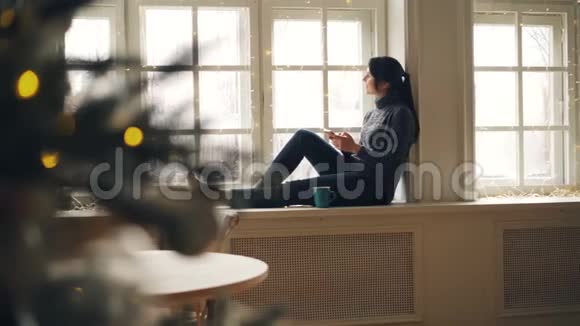 新年快乐的女孩和坐在窗台上的朋友交流用智能手机触摸屏幕视频的预览图