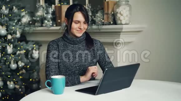 穿着毛衣的微笑着的年轻女士正在圣诞节时坐在新年树附近思考和打字现代视频的预览图