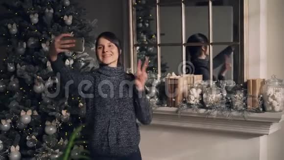 圣诞当天美丽的年轻女士正站在装饰好的壁炉和新年旁用智能手机自拍视频的预览图