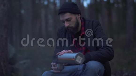 小胡子坐在篝火旁的森林里把饮料从热水瓶里放进杯子里喝无胡子的家伙视频的预览图