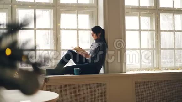在圣诞节那天一个放松的女孩正在看书她坐在窗台上12月穿着衣服看着窗外视频的预览图