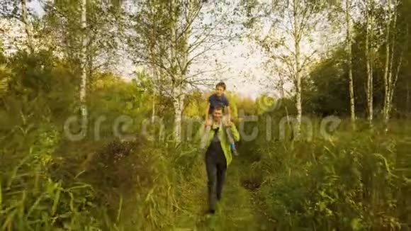 父亲背着儿子在夏天日落前肩上扛着儿子穿过森林和高高的草地慢动作视频的预览图