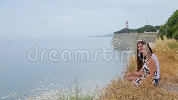 积极向上的金发女孩和黑发女孩坐在悬崖边微笑着看着无边无际的蓝色大海视频的预览图