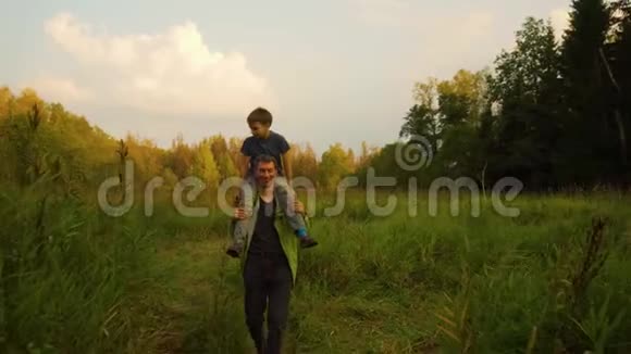 父亲背着儿子在夏天日落前肩上扛着儿子穿过森林和高高的草地慢动作视频的预览图