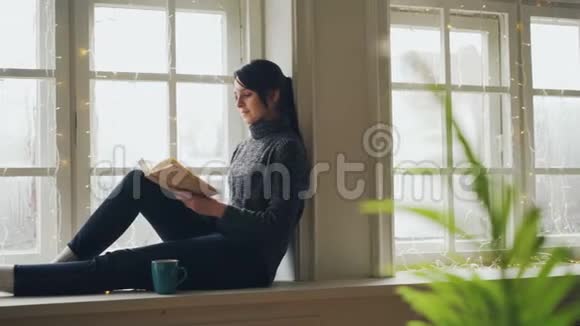 穿着毛衣和牛仔裤的漂亮黑发女人正在看书在圣诞节的时候坐在窗台上享受着免费的孤独视频的预览图
