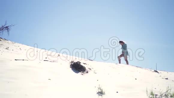 年轻女子在蓝天背景下的炎热沙漠中的沙地沙丘上行走美丽的女孩走在沙滩上视频的预览图