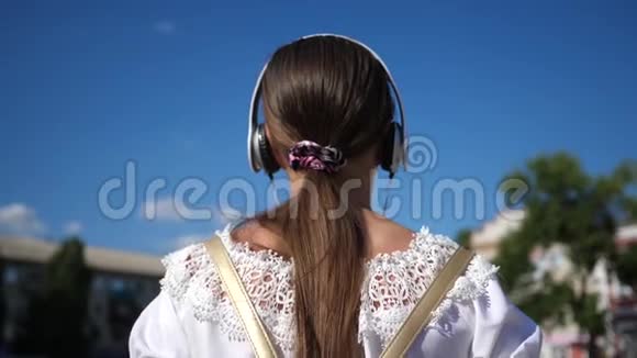 一个年轻的女孩带着耳机走在城市的街道上听音乐穿着白色连衣裙留着长发的少女视频的预览图