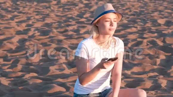 美丽的金发女孩戴着帽子坐在海滩上日落时用手机视频的预览图