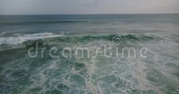 无人机倾斜下来跟随蓝色的大海浪到达海岸并在阴天与白色海泡形成碰撞视频的预览图