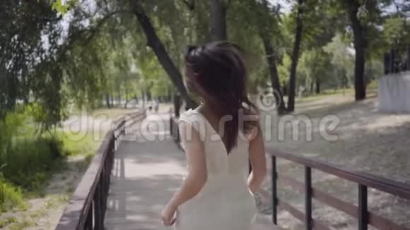 可爱的年轻女孩一头黑发戴着太阳镜穿着白色的夏季时装长裙穿在木桥上视频的预览图
