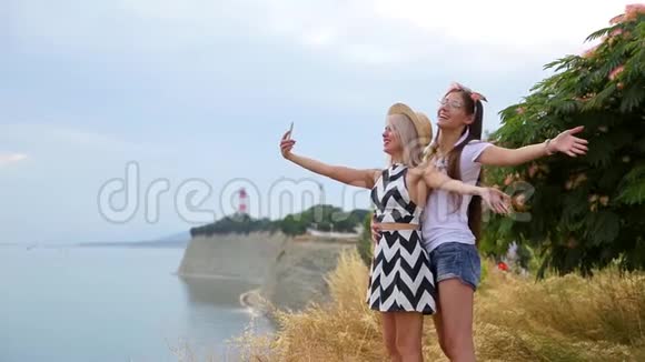 可爱的积极的金发女孩和黑发女孩唱着歌用求助电话在靠近岩石悬崖的海边射击视频的预览图