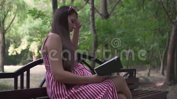 一个怀孕的女孩坐在公园的长凳上一个穿着条纹白红色连衣裙的女孩热情地看书视频的预览图