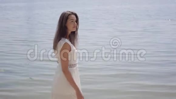肖像迷人的年轻女孩留着长长的黑发穿着一件白色的夏季时装长裙站在河边视频的预览图