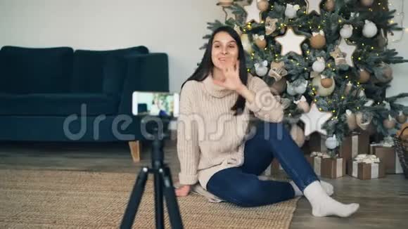 情绪激动的年轻女子坐在圣诞树附近的地板上用智能手机为网上博客录制视频视频的预览图